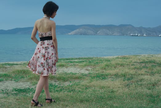 A young woman in a floral dress gazes at the calm sea with mountains in the background.