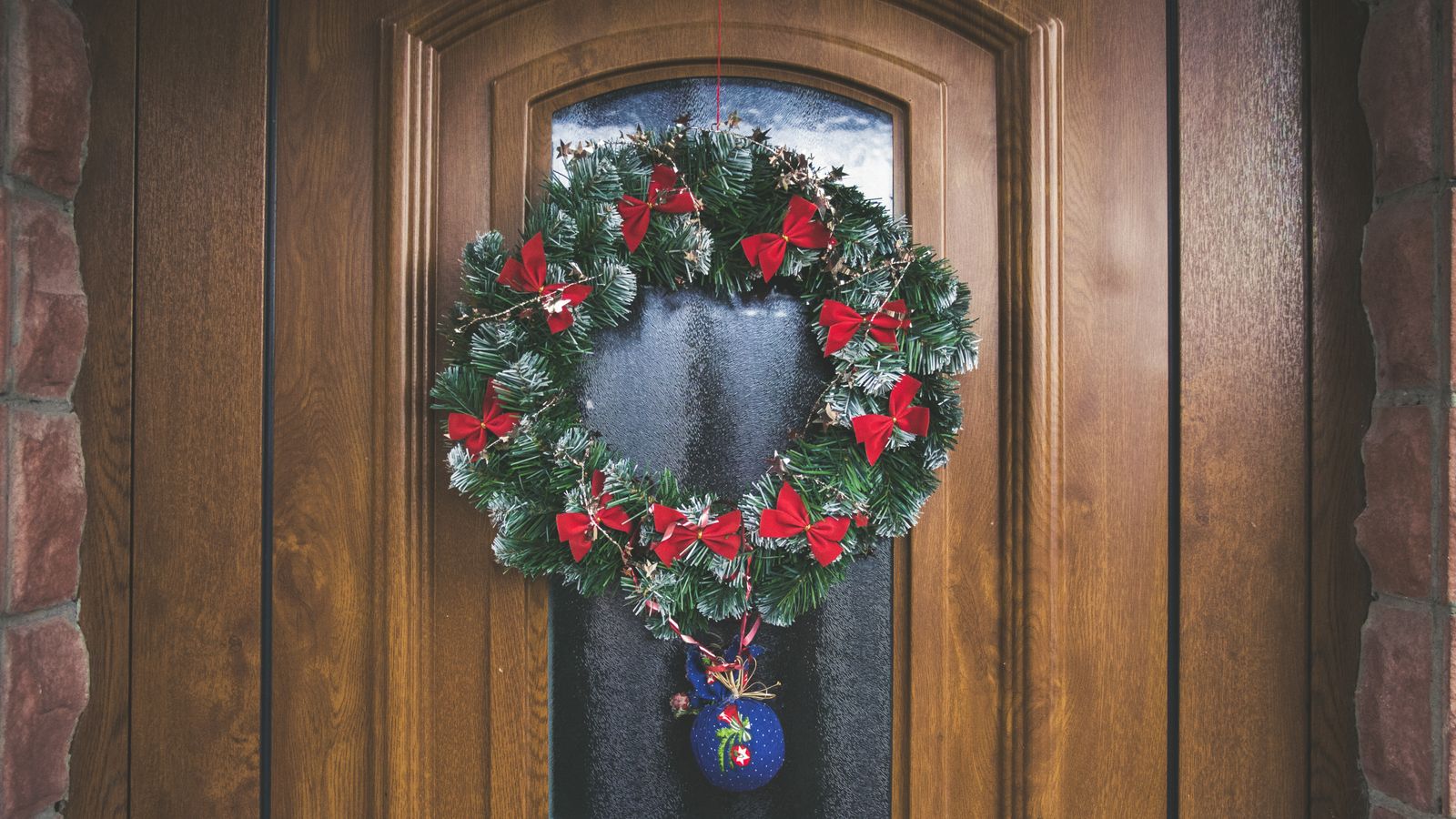 An evergreen Christmas wreath with red bows and a hanging ornament on a wooden front door.