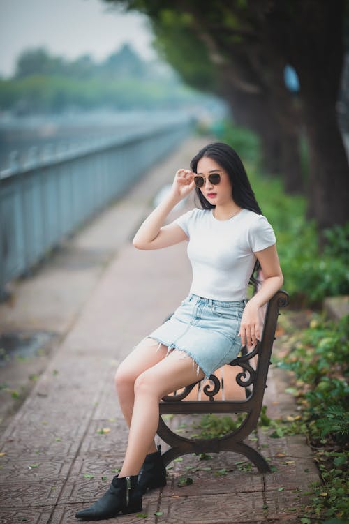 Trendy young ethnic lady adjusting sunglasses on bench in park