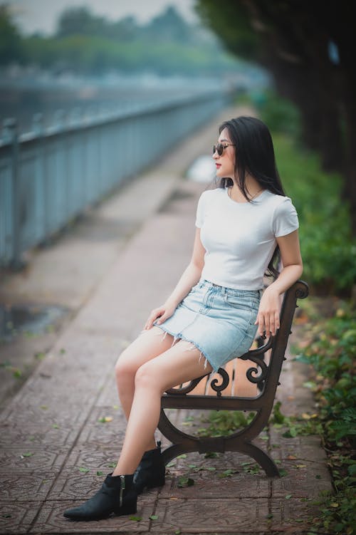 Full length of stylish young ethnic lady with long dark hair in trendy outfit and sunglasses sitting on bench in city park and looking away