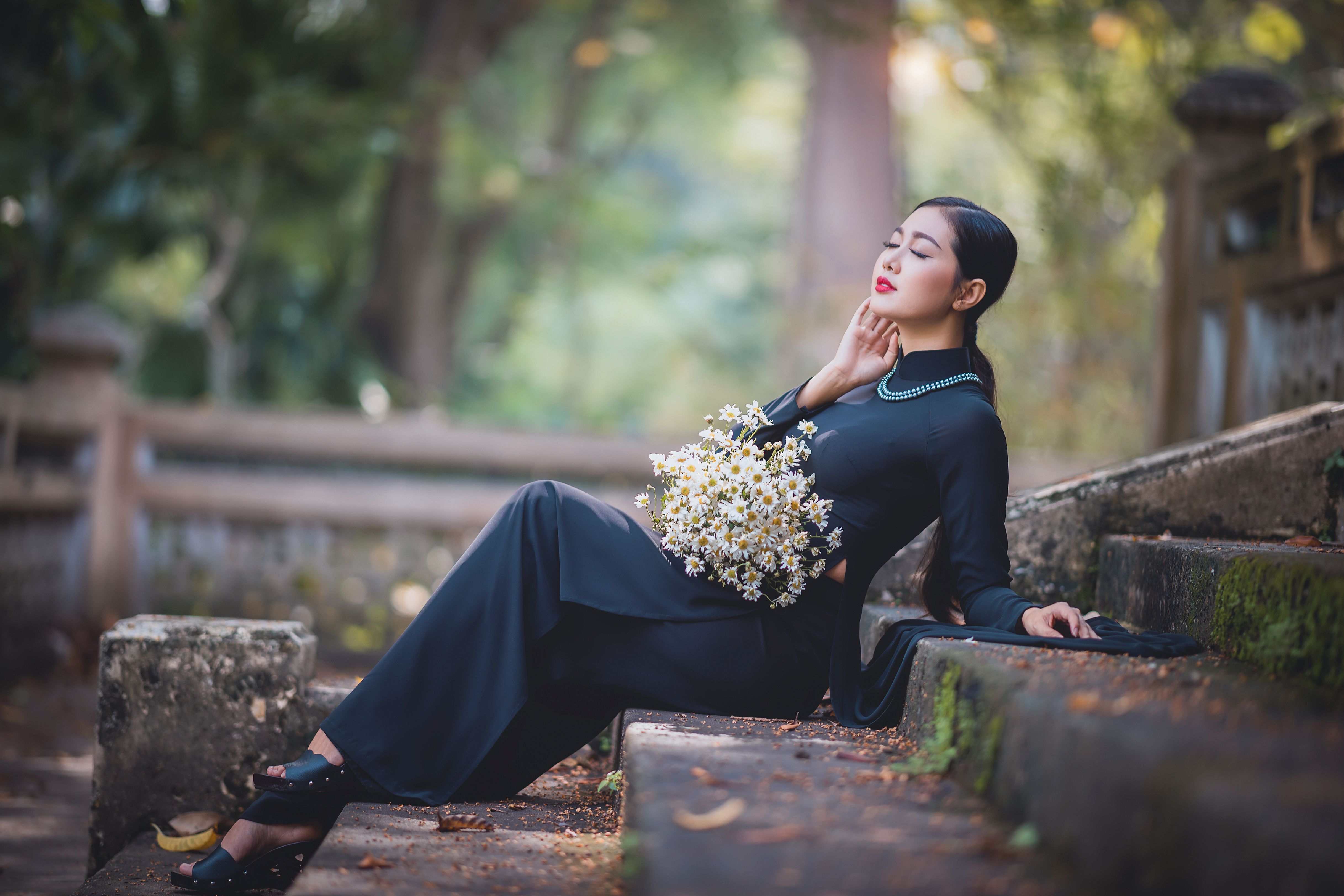 Side view of relaxed young ethnic woman in elegant dress touching face with closed eyes while sitting on stone stair in autumn park with bunch of fresh chamomiles
