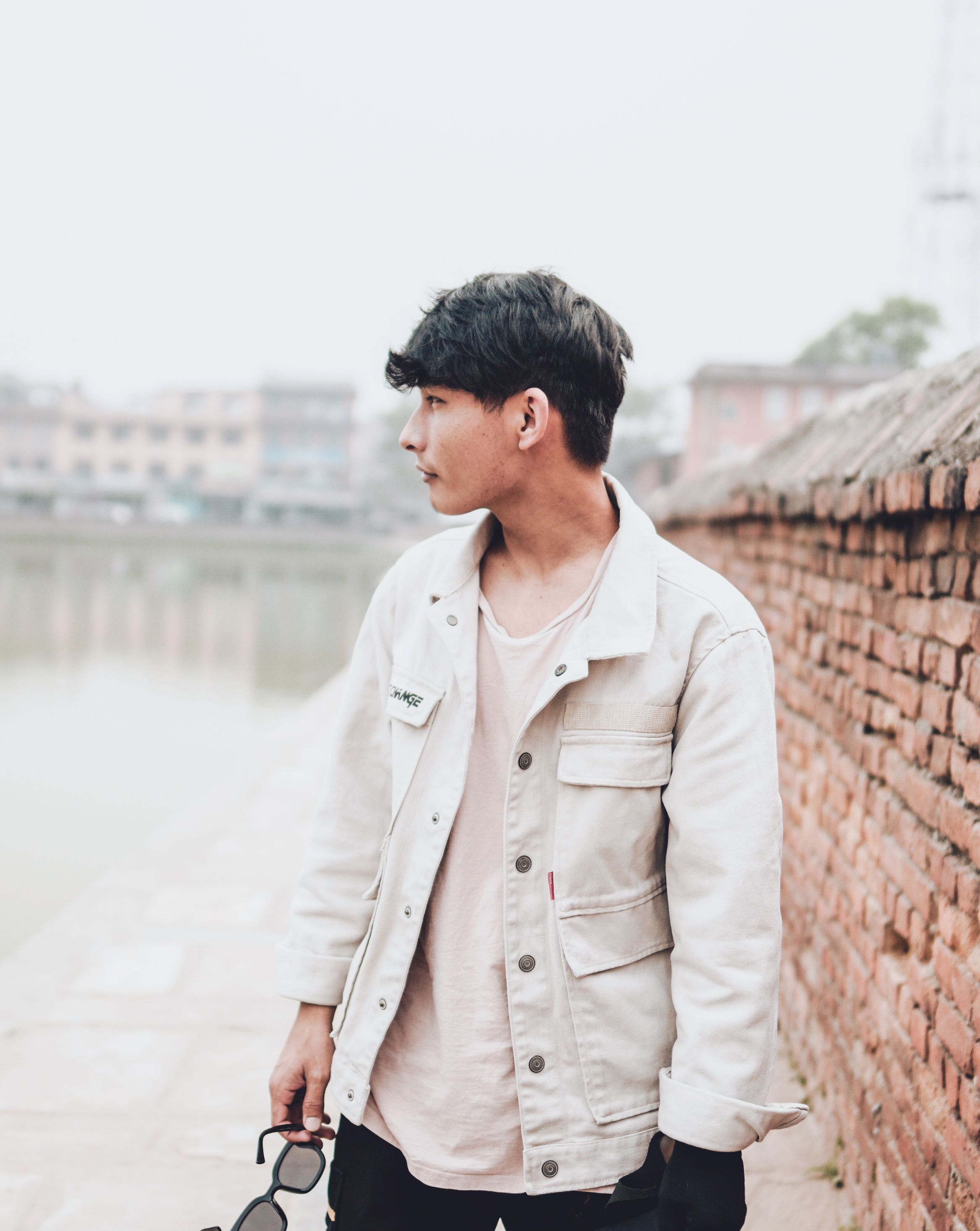 Young man holding sunglasses by a brick wall, looking away thoughtfully outdoors.