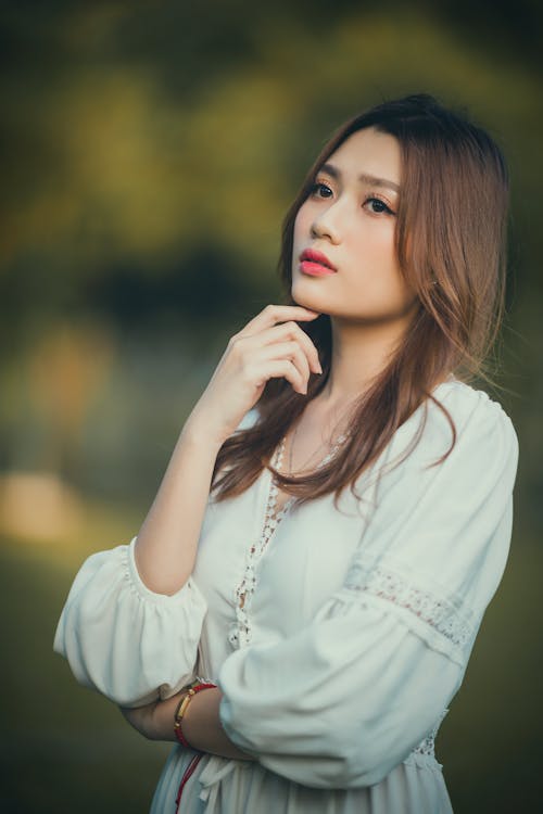 Thoughtful young elegant Asian lady touching chin and looking away in nature