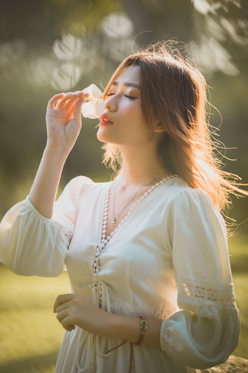 Romantic young Asian female with long brown hair in elegant white dress enjoying smell of gentle flower with closed eyes while standing on grassy meadow in nature on sunny day