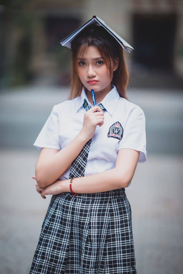 Unhappy Ethnic Teenage Female Student Standing On Street With Copybook On Head