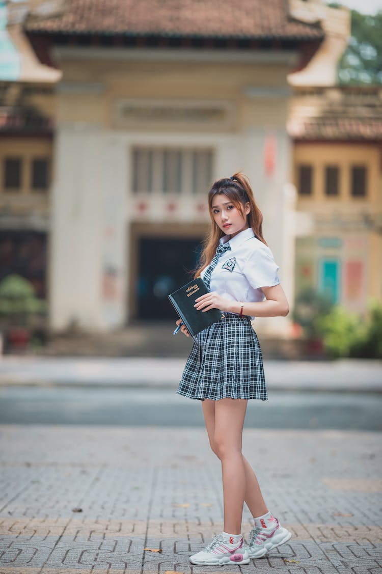 Asian Schoolgirl In Uniform With Book Near Building In Street