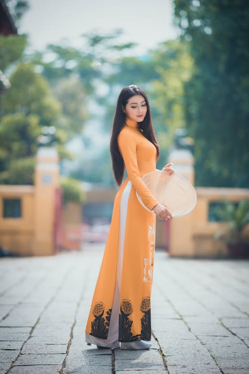 Side view of pensive Asian woman with long dark hair in stylish dress standing with conical hat in hands on pavement walkway against green trees in daytime