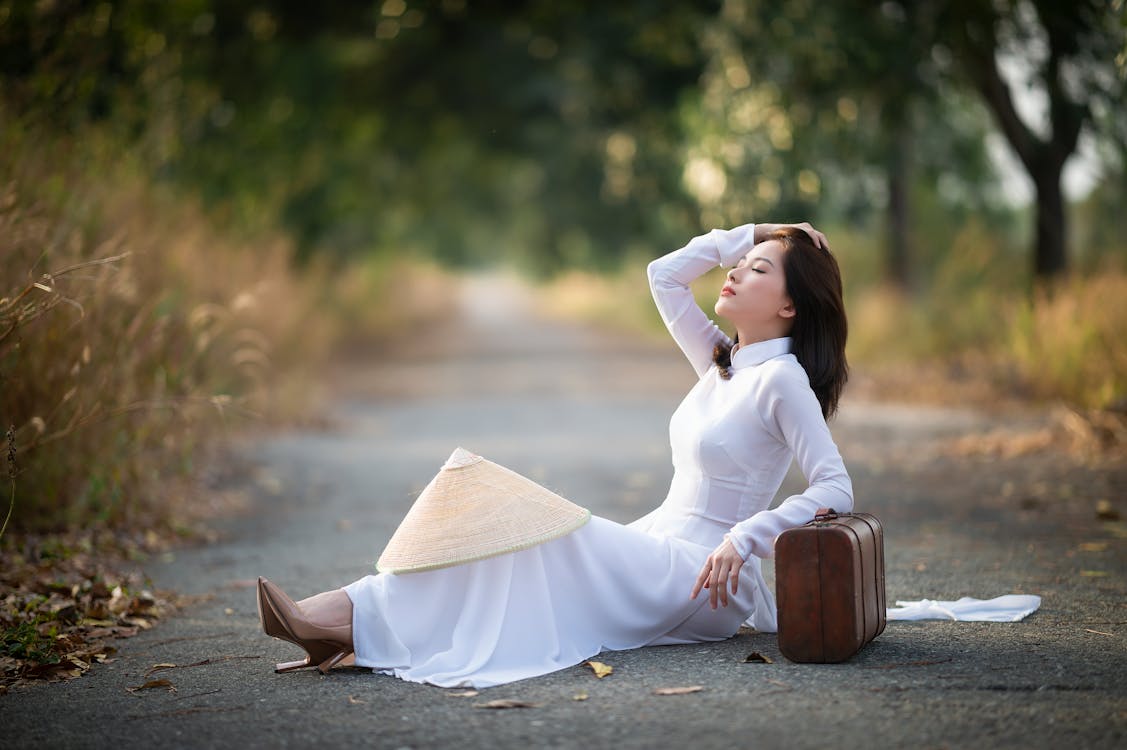 Side view of calm Asian female in white dress sitting with closed eyes on asphalt walkway and leaning on suitcase in park in daytime