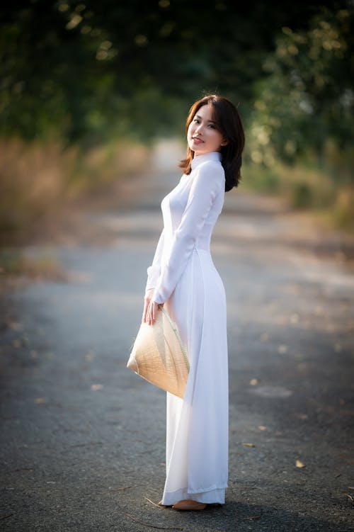 Side view of charming young Asian woman in long white dress standing with conical hat in hands and looking at camera on asphalt walkway in park in daylight