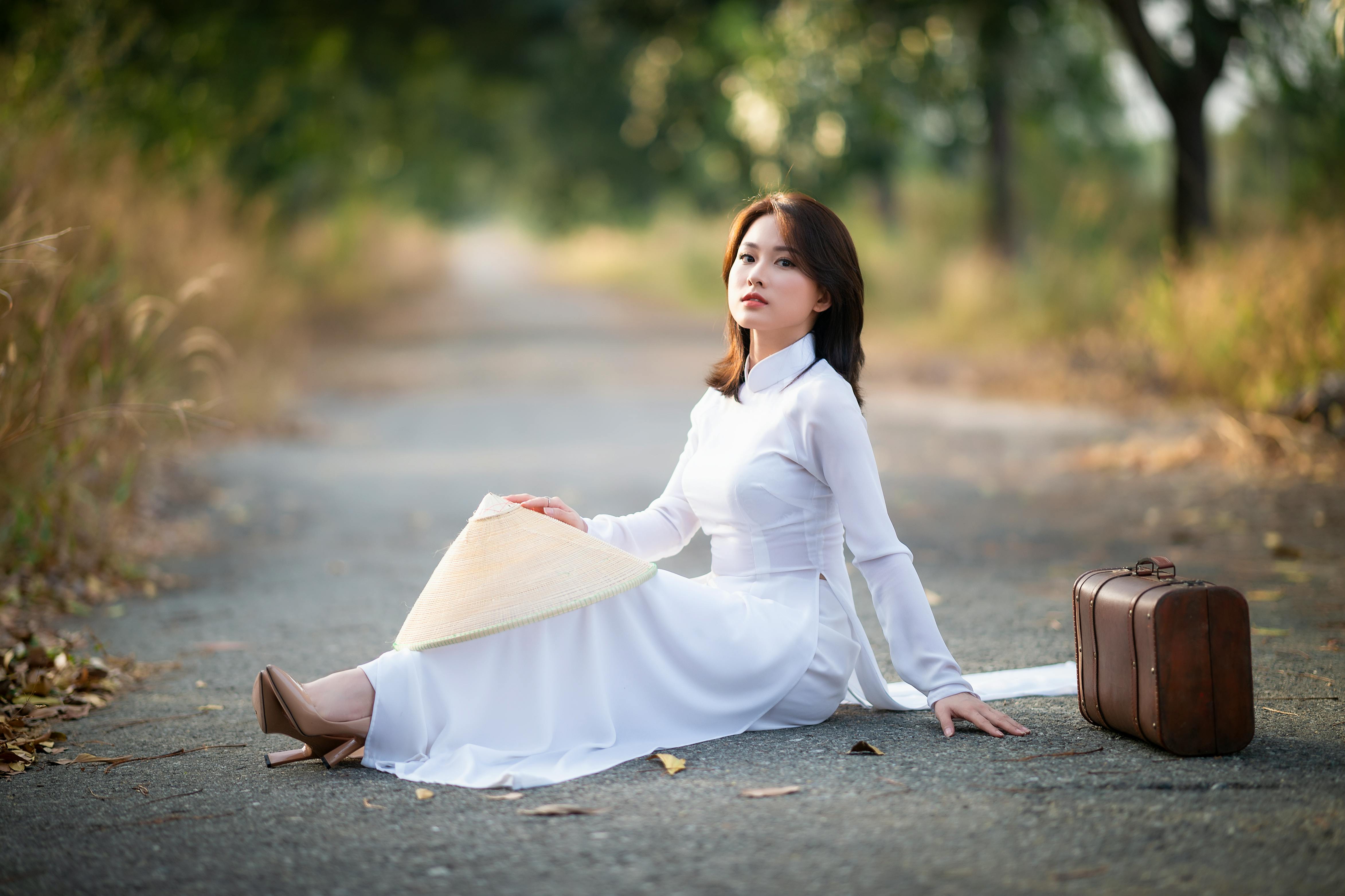 Free Asian woman in a white dress with a conical hat and suitcase beside her on a rural road. Stock Photo