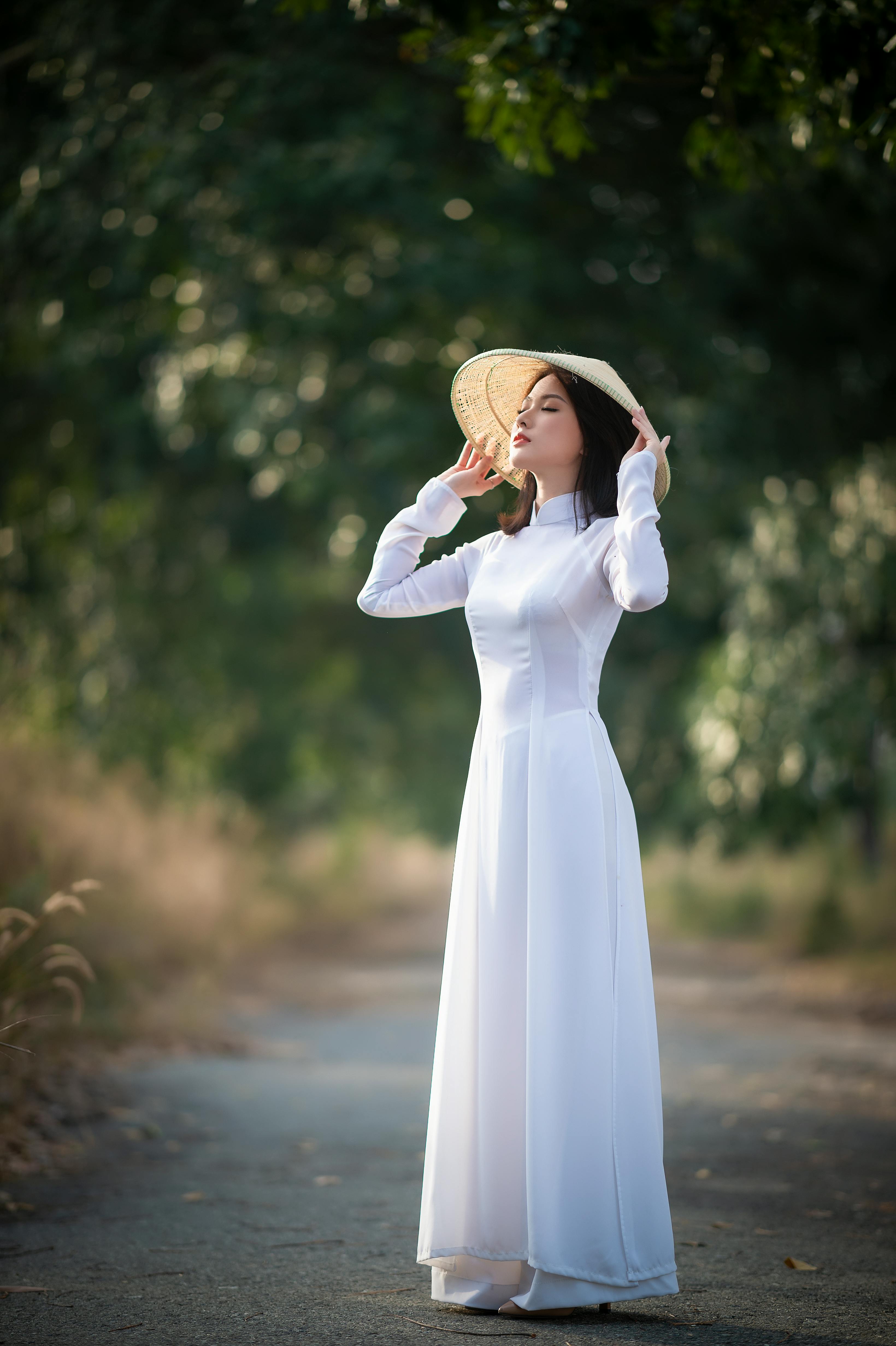 Free Elegant Asian woman in a white dress and conical hat posing in a serene outdoor setting. Stock Photo