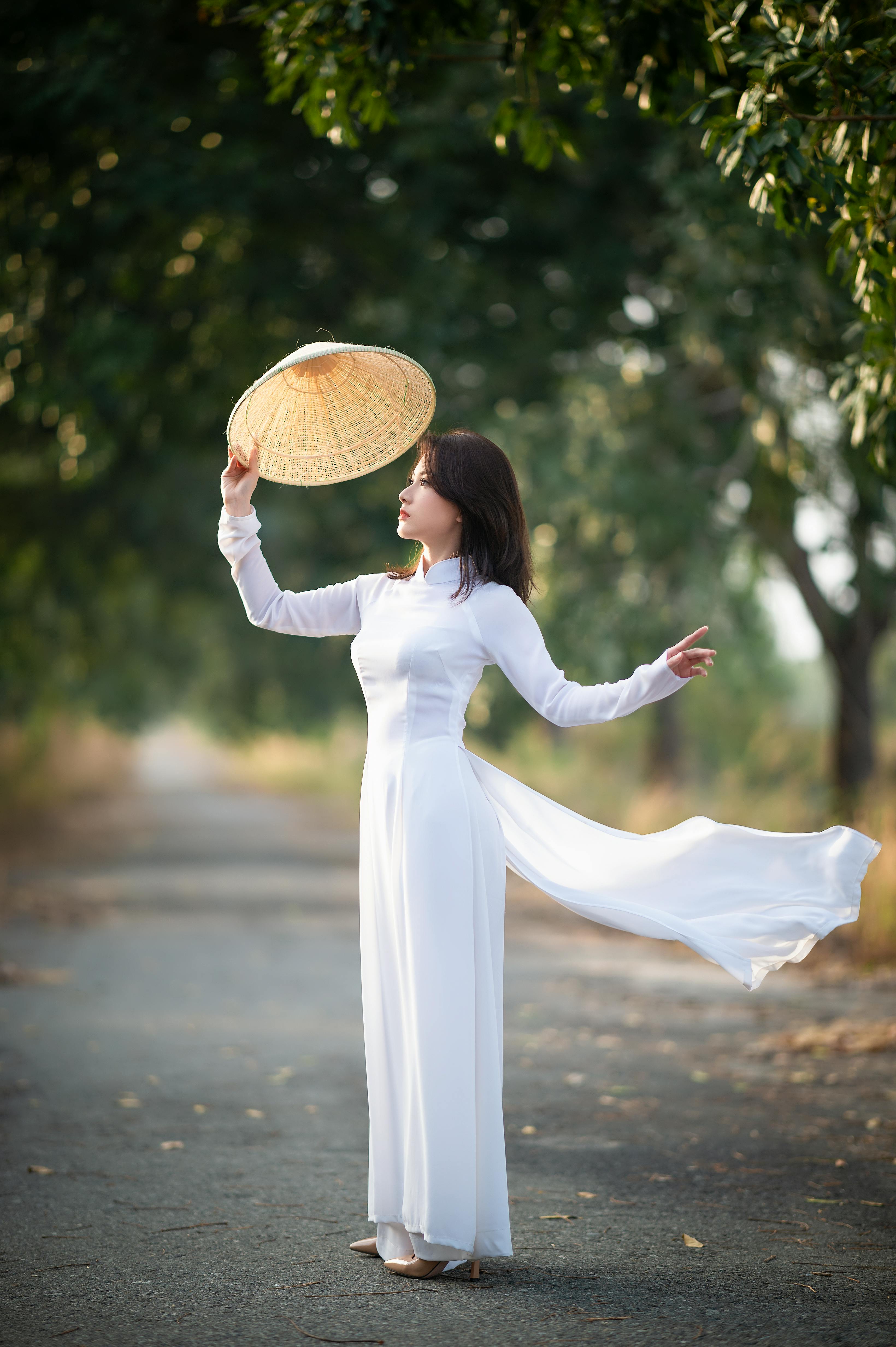 Free An elegant woman in a flowing white dress poses on a tree-lined road with a conical hat. Stock Photo