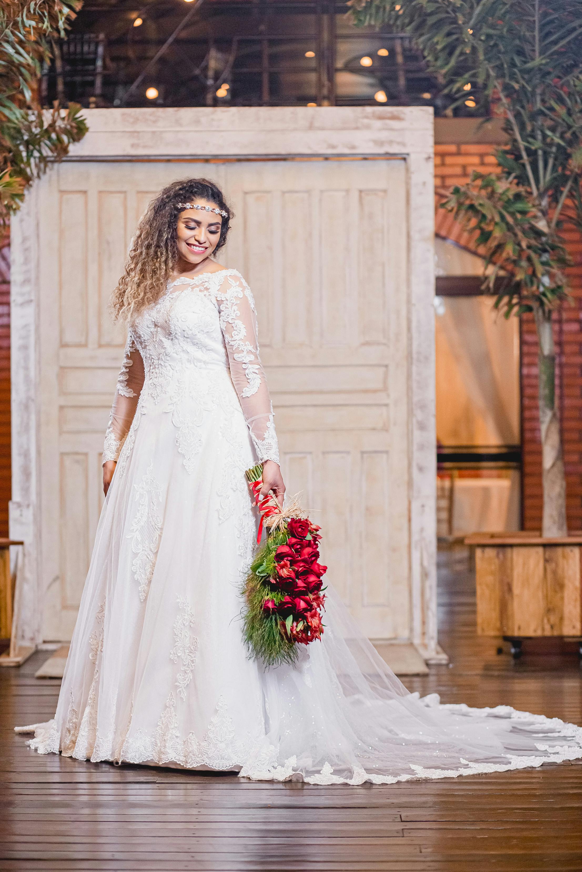 Beautiful bride in lace gown holding vibrant red bouquet indoors with rustic decor.
