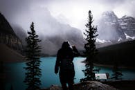 冒险家身着深色服装，凝视着云雾缭绕的冰沙湖，周围高耸的松树和崎岖的山峰环绕