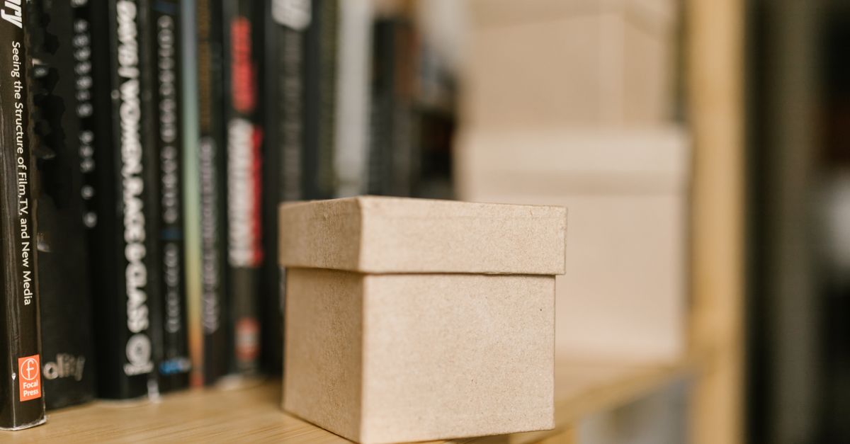 Foto de close-up de uma pequena caixa de papel marrom em uma prateleira de madeira com livros ao fundo.