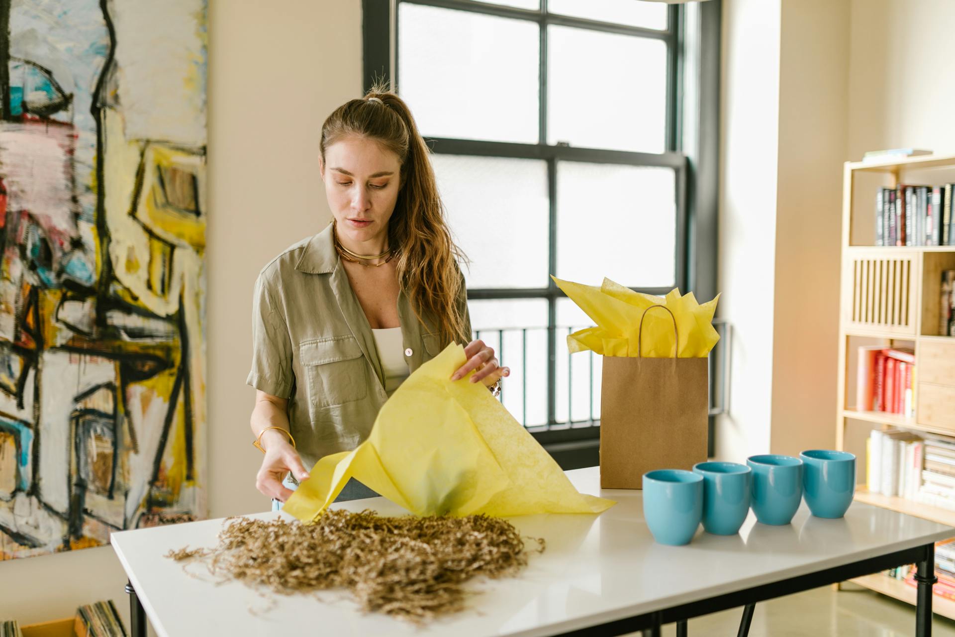A woman in a green shirt wraps gifts in yellow tissue paper at a work table.