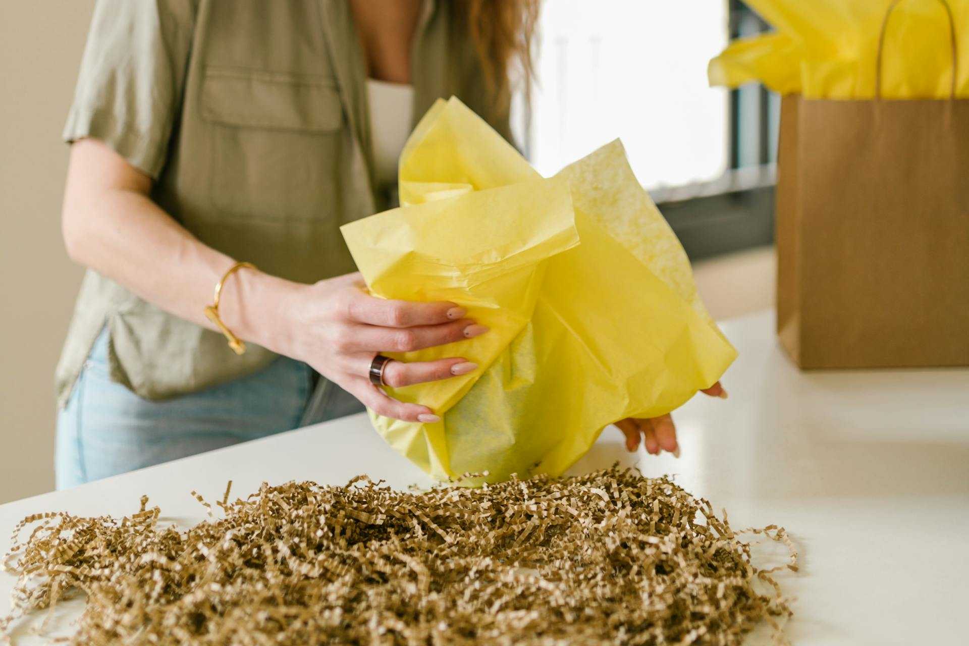 A person wrapping a gift with bright yellow tissue paper and shredded paper on a table.
