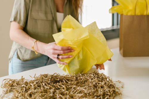 Free A person wrapping a gift with bright yellow tissue paper and shredded paper on a table. Stock Photo