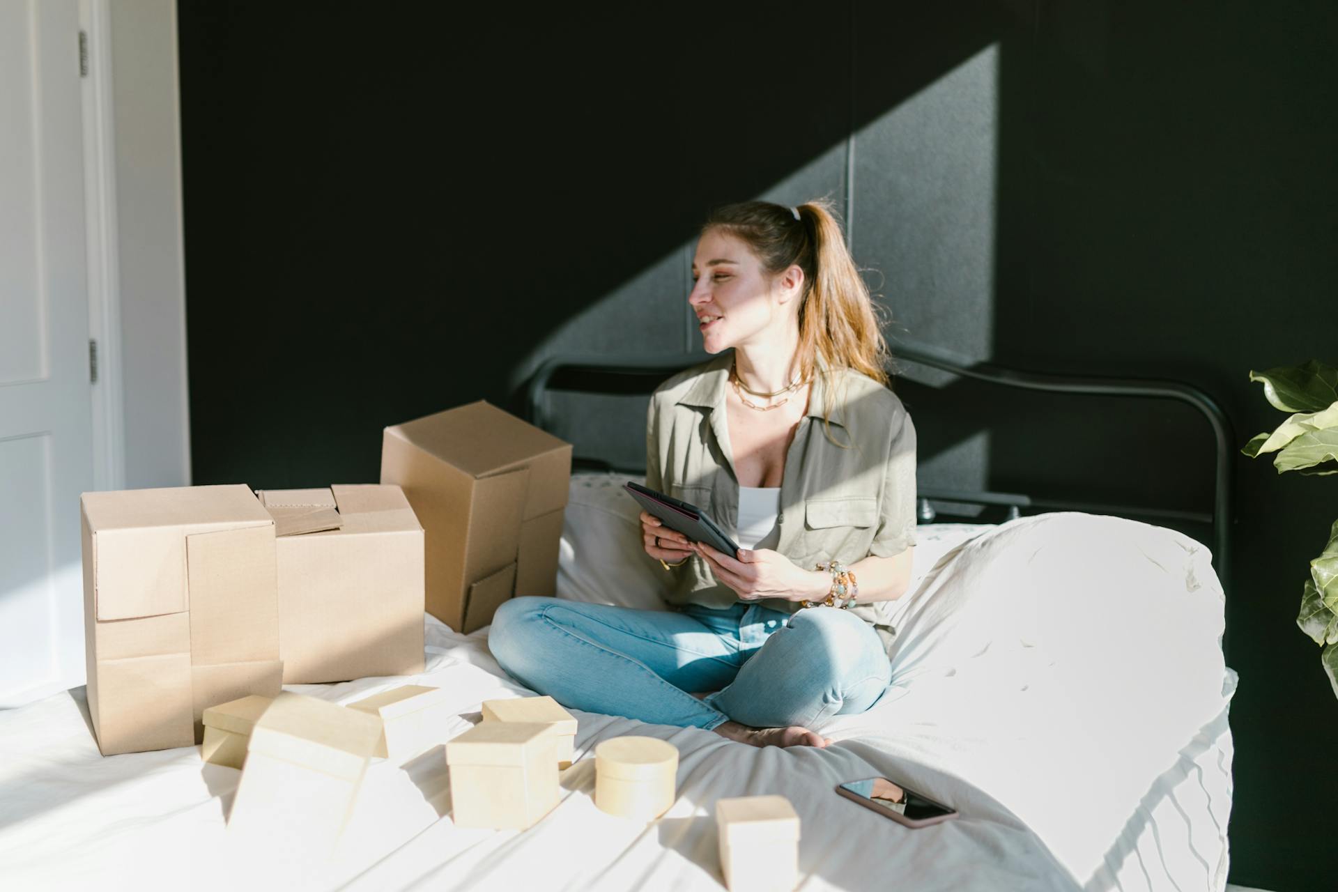 Woman sitting on bed with boxes and tablet, running an online business.