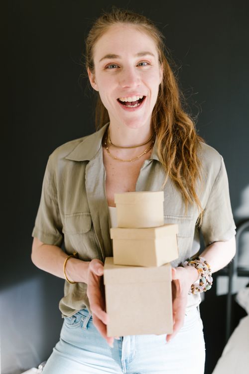 Free A cheerful young woman holding small cardboard boxes indoors, wearing a casual outfit. Stock Photo