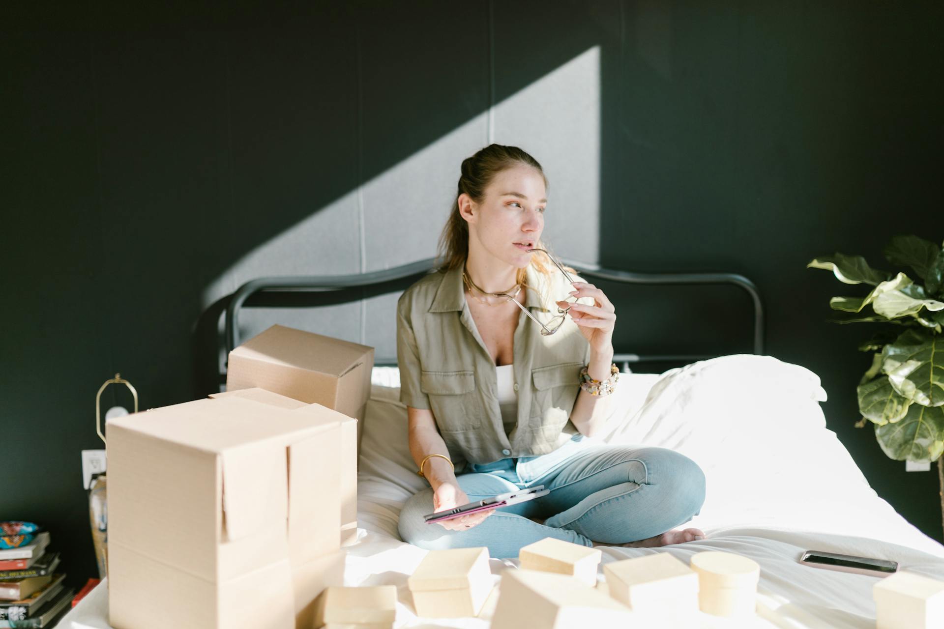 A woman sits with tablet and coffee, organizing her home-based e-commerce setup with boxes.