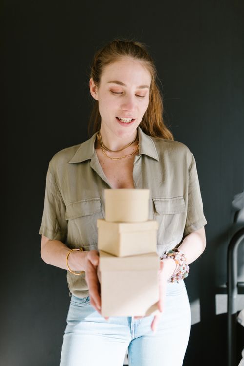 Free Cheerful woman holding stacked cardboard boxes in a bright indoor setting, symbolizing small business entrepreneurship. Stock Photo