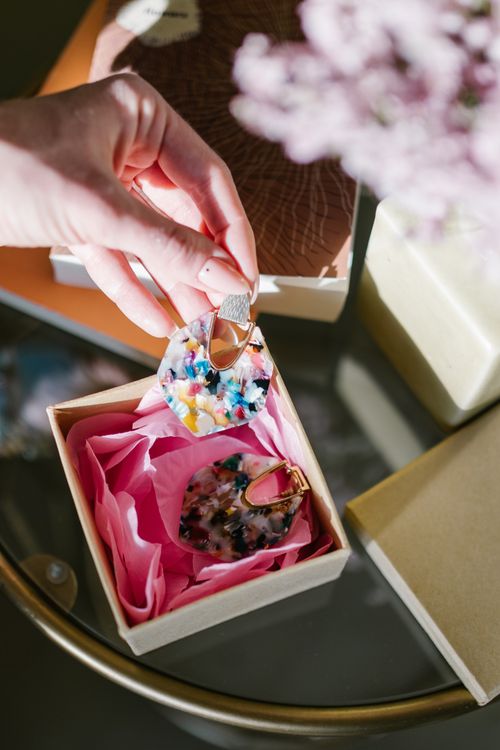 Free Close-up of a hand holding vibrant earrings above a gift box, adorned with pink tissue paper. Stock Photo