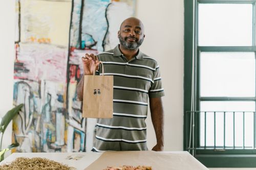 Free A smiling entrepreneur holding a paper bag with stickers in an artistic setting, showcasing small business creativity. Stock Photo