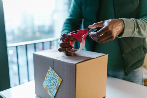 Free Close-up of man sealing a cardboard box with a tape dispenser indoors. Stock Photo