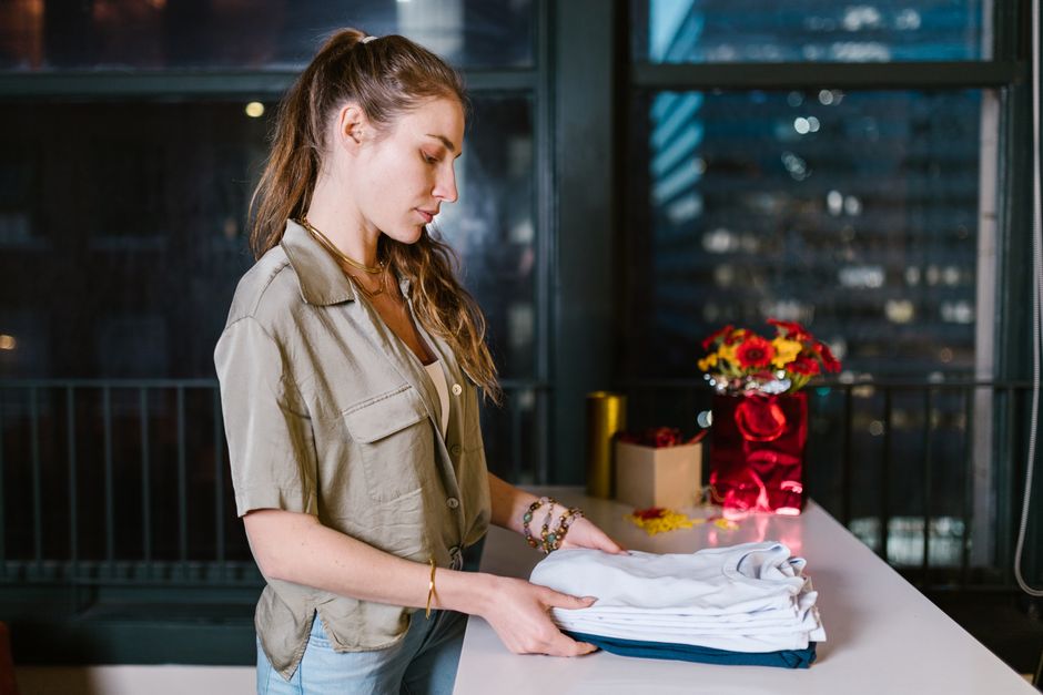 Caucasian woman folding shirts at a table indoors, with city lights in the background.