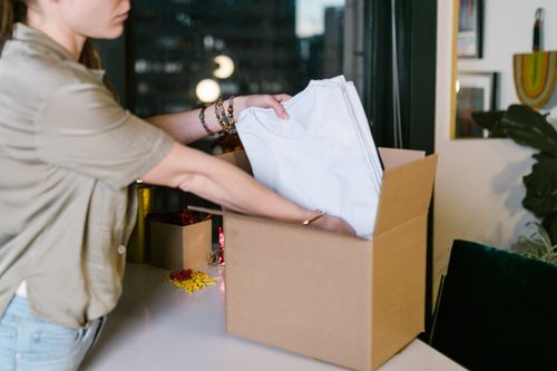 Free A woman packs white t-shirts into a cardboard box, set in a modern indoor environment. Stock Photo