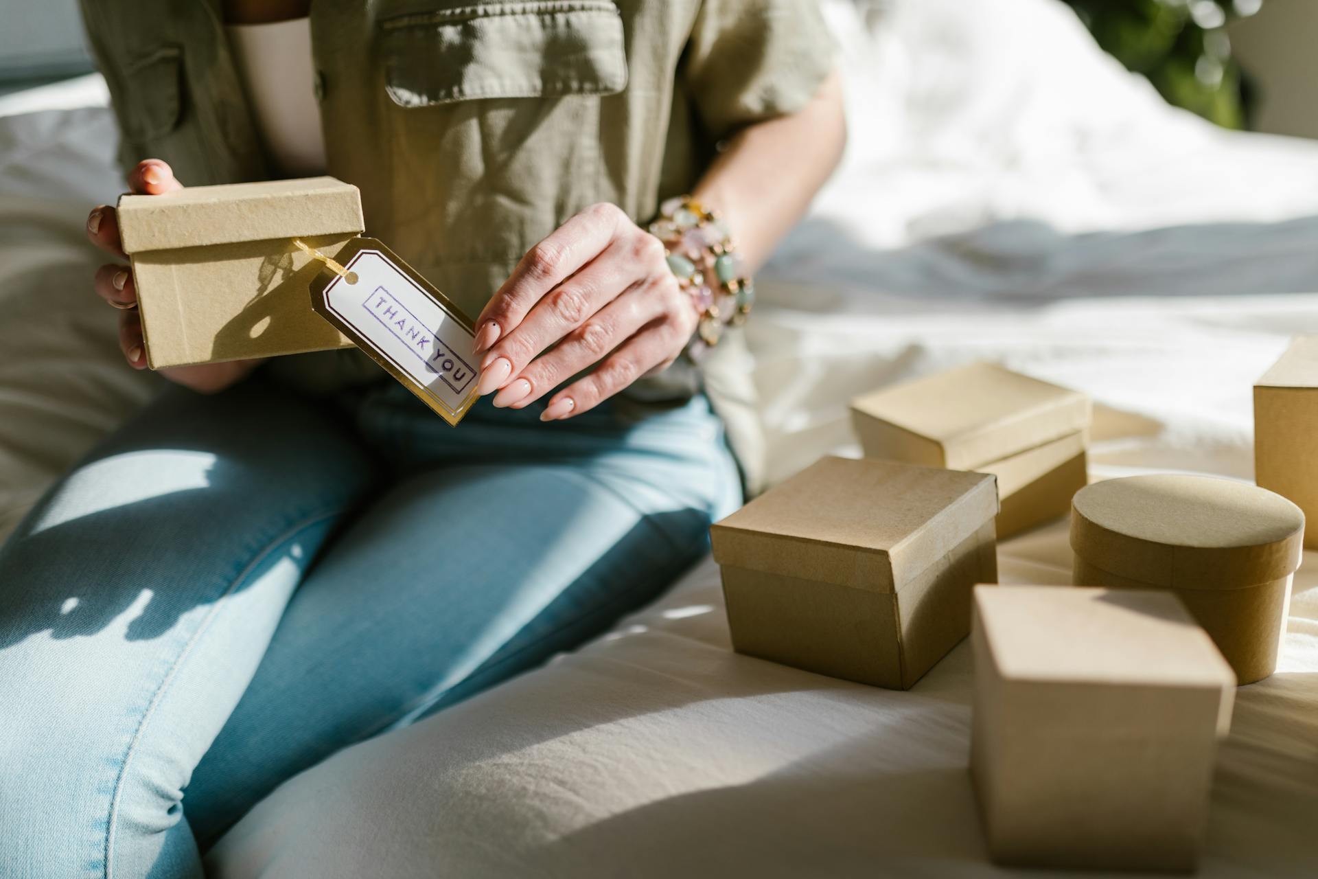 Close-up of a woman holding a thank you gift box surrounded by cardboard boxes on a bed.