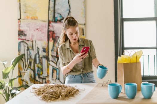 Young woman photographing blue mug in a creative art studio setting, surrounded by supplies.