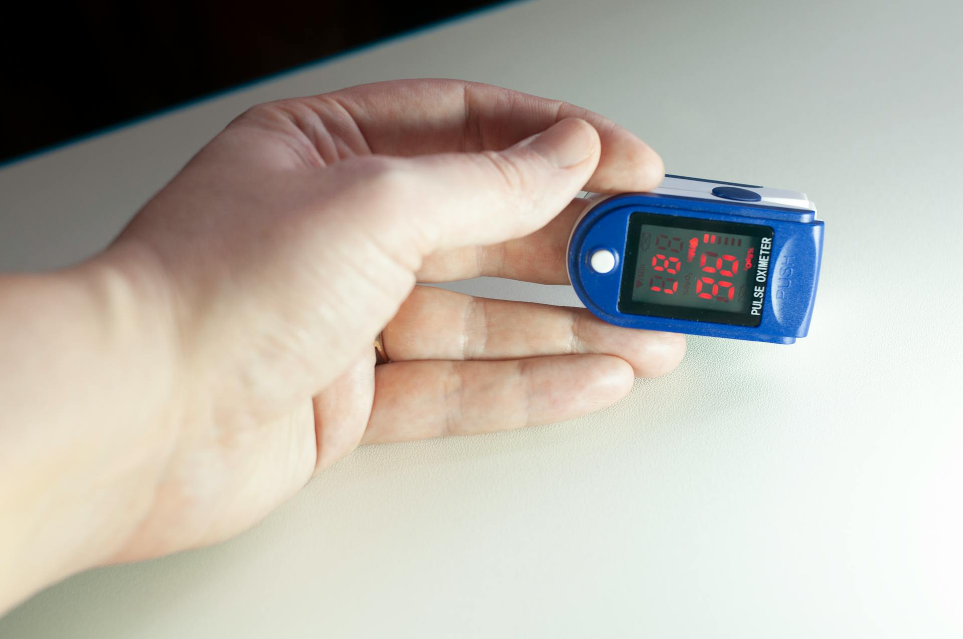 A hand checking oxygen saturation with a pulse oximeter device on a light surface.