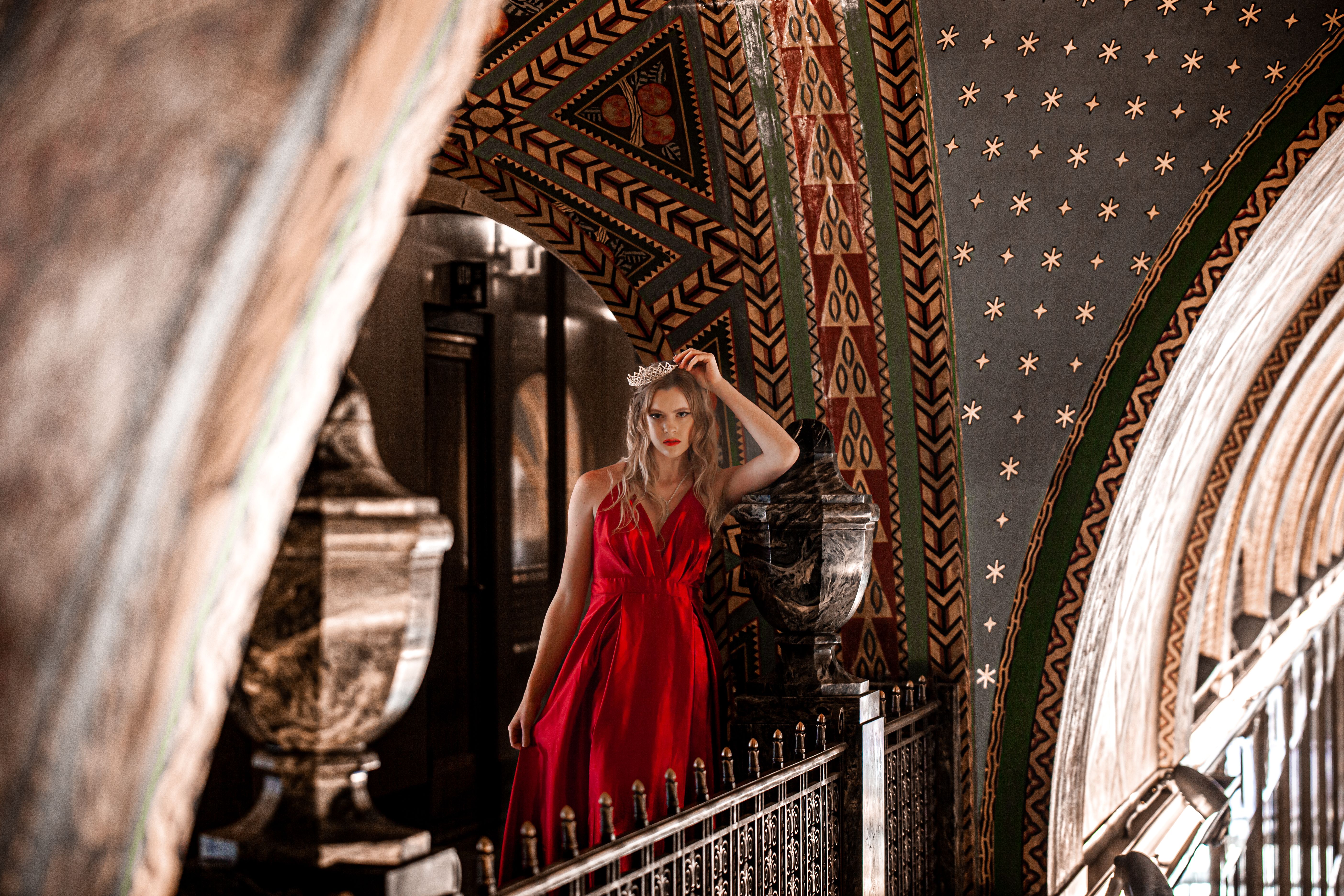 Stunning woman in a red gown poses elegantly under an ornate archway, showcasing fashion and architecture.