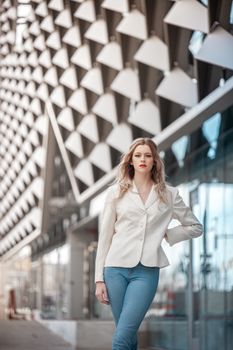 Fashionable woman in white blazer and jeans poses outdoors in modern cityscape.