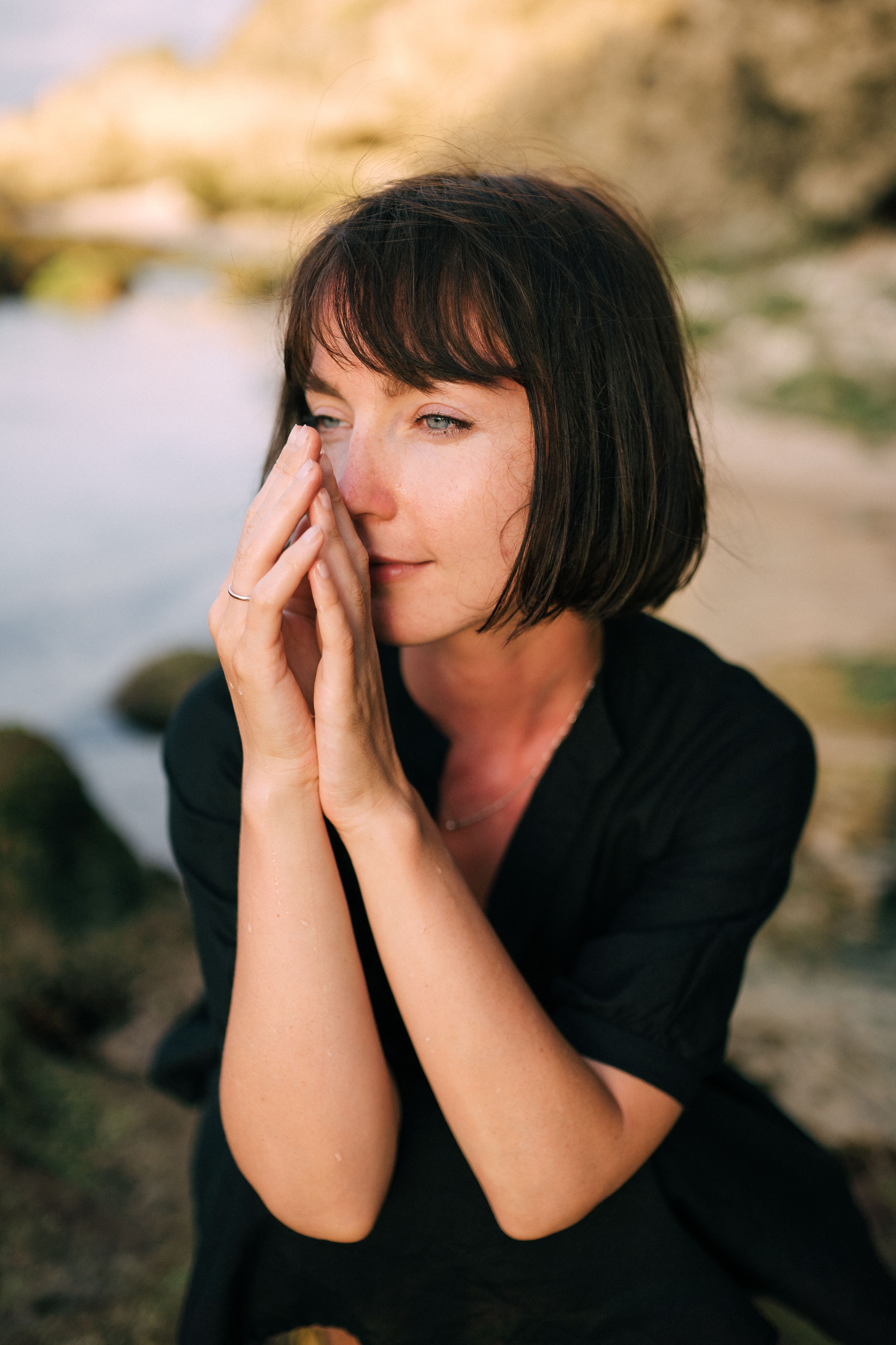 A thoughtful woman with short hair gazes into the distance at a serene beach setting, hands together in contemplation.