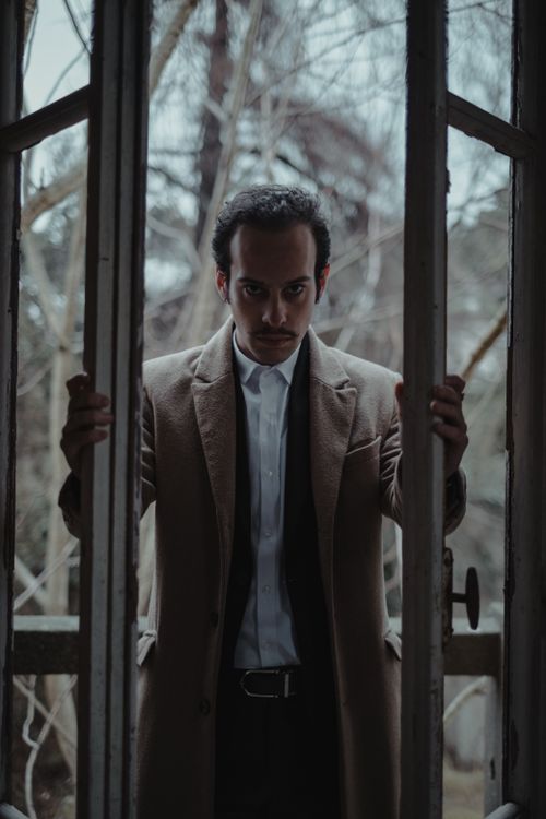 Free A serious man with a mustache stands in a coat by an open glass door, looking outdoors. Stock Photo