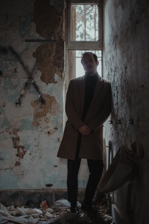 Free A man in a coat stands in a dimly lit, abandoned room with peeling walls. Stock Photo