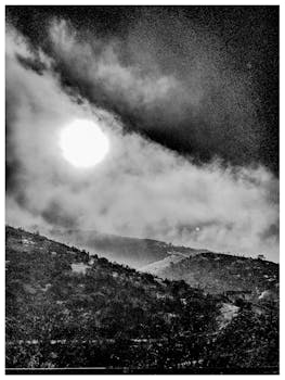 Moody black and white photo of mountains with a cloudy sky and visible moon.