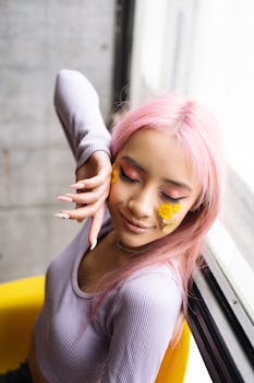 Young woman with floral creative makeup and pink hair smiling indoors by the window.