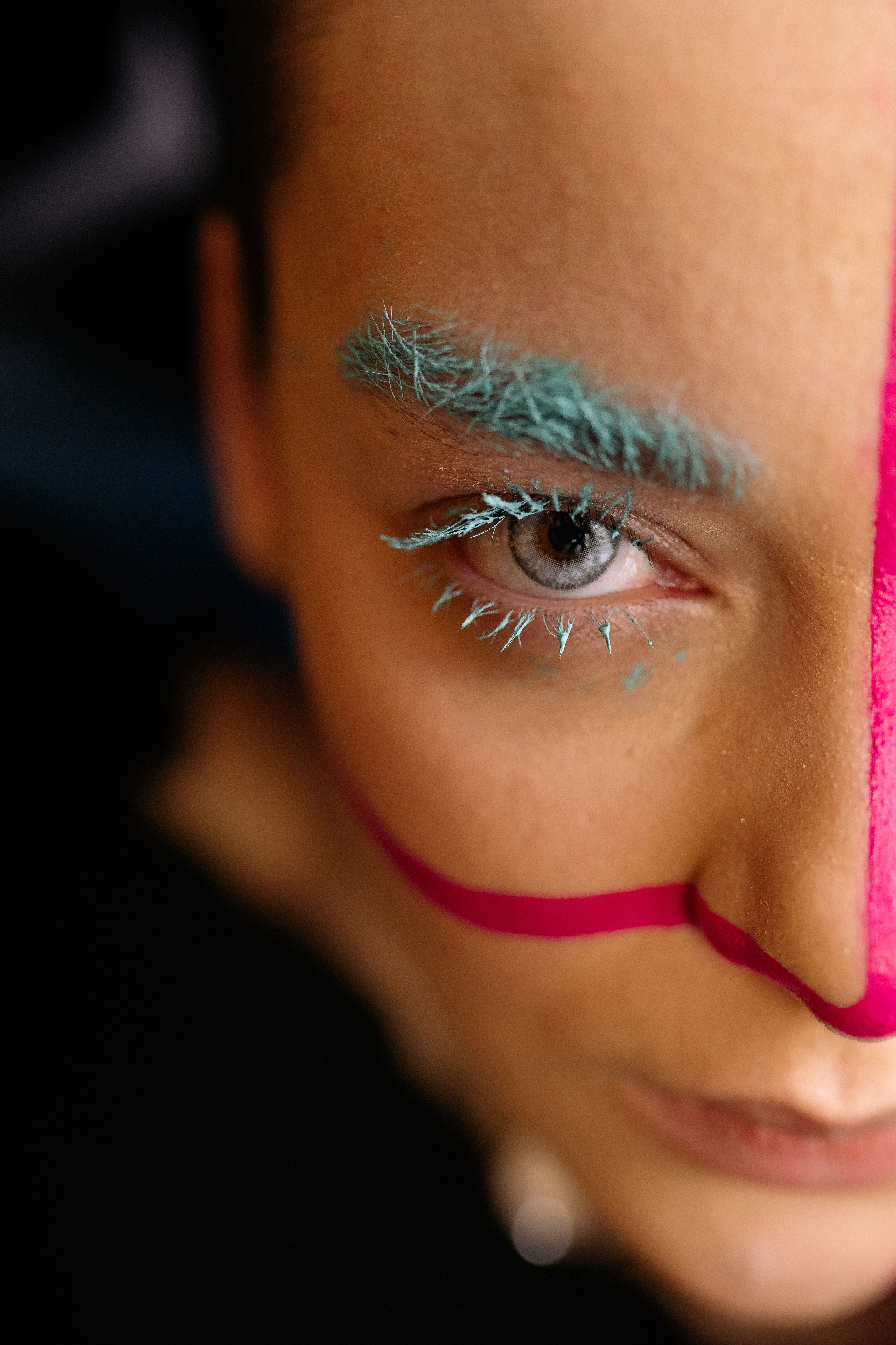 Retrato em close de uma pessoa com maquiagem colorida apresentando cílios e sobrancelhas azuis, e pintura facial rosa.