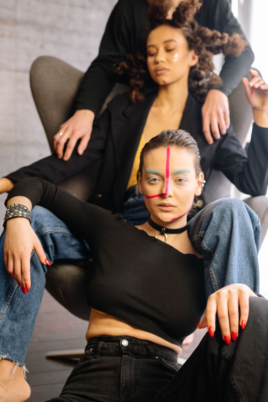 Stylish women in black clothing with colorful makeup poses in a modern artistic setting.
