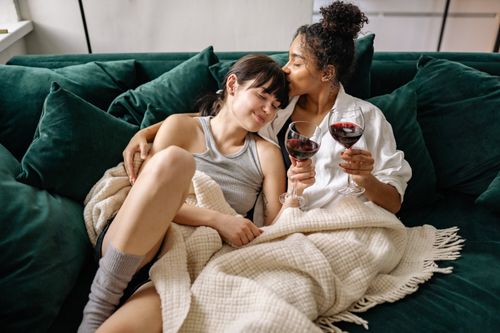 Free A loving lesbian couple snuggled on the couch with glasses of red wine, embracing warmth and affection. Stock Photo