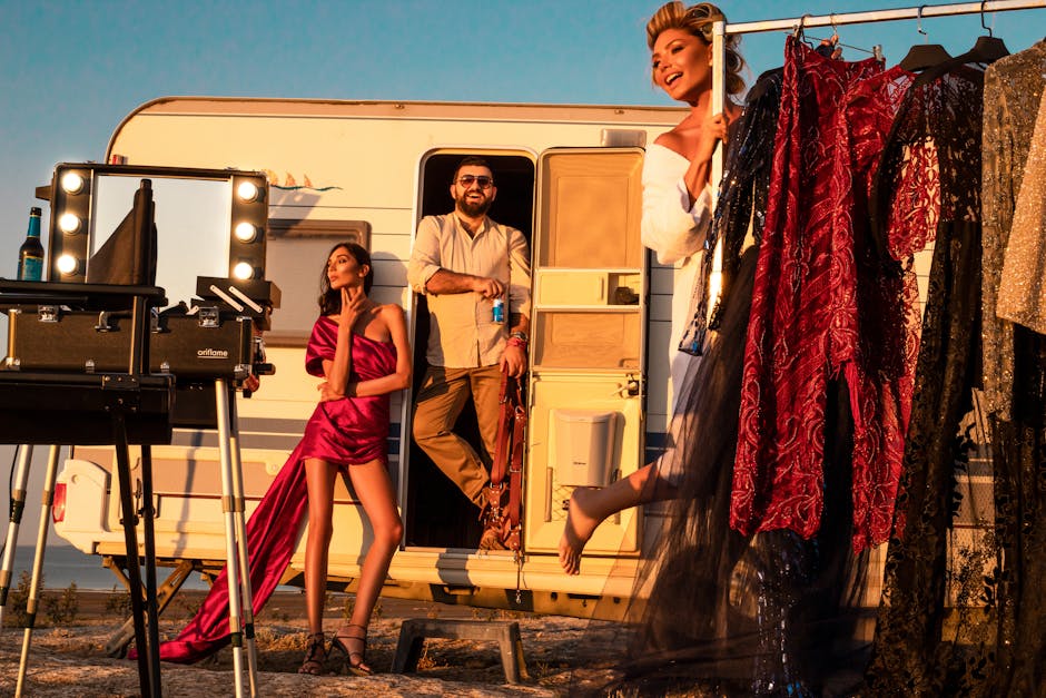 Full body of cheerful male standing in trailer near gorgeous women wearing stylish dresses on street with collection of outfits on rack and professional supply while preparing for video shoot