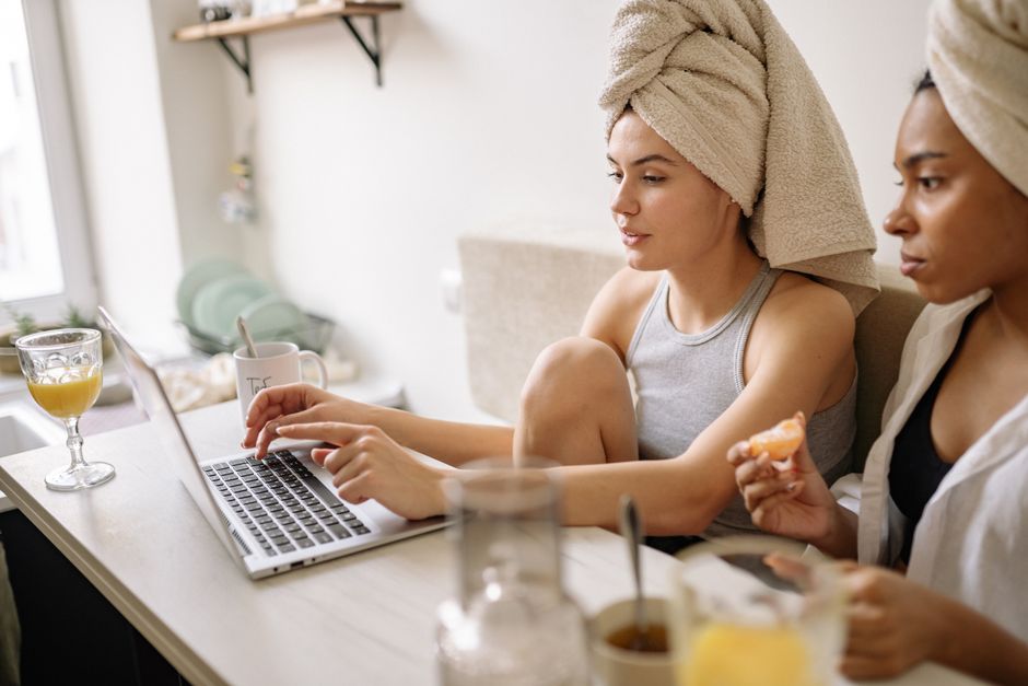 Two women in towels use a laptop at home, enjoying snacks and drinks in a cozy setting.