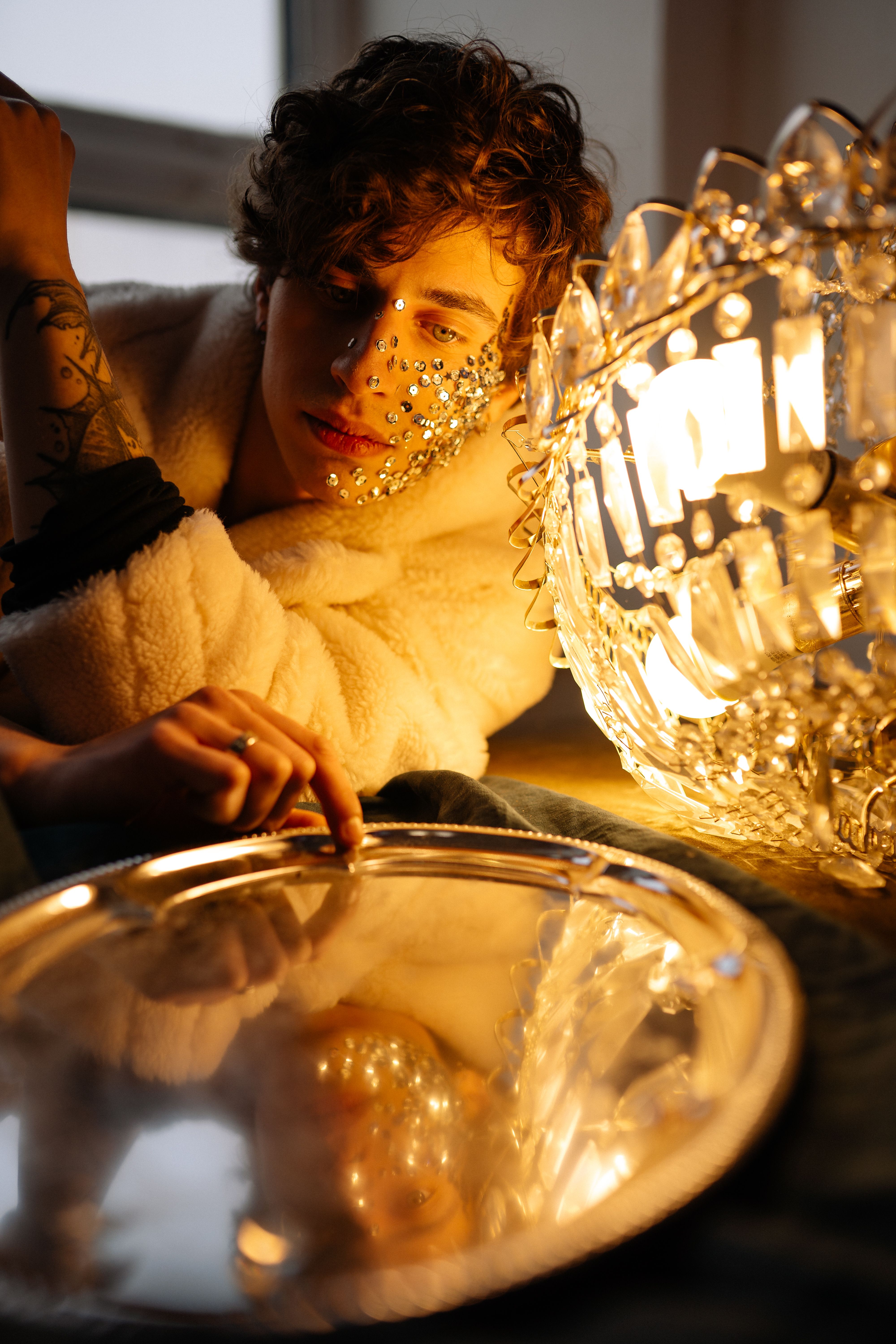 A young model with art makeup poses beside an illuminated chandelier indoors.