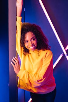 A young woman smiling with art makeup in neon lights, wearing a yellow sweater, embodying modern and vibrant fashion.