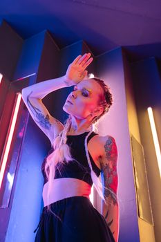 A young tattooed woman in a black crop top poses with dramatic neon lighting in an artistic portrait.