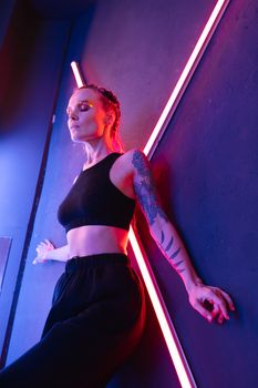 Fashionable young woman posing stylishly under neon lights with glitter makeup and tattoos.